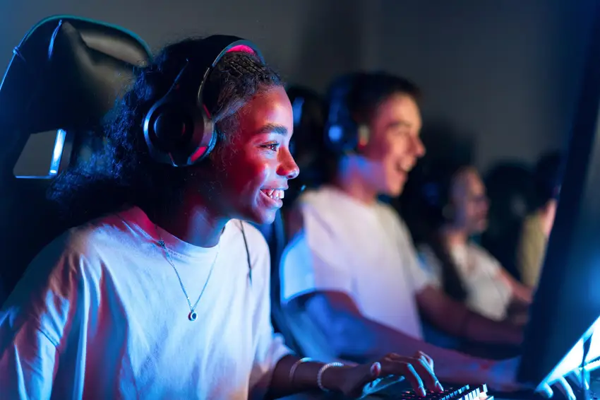 Een vrouwelijke gamer met een headset op, die glimlachend op een computer speelt terwijl haar teamgenoten op de achtergrond te zien zijn; ze geniet van een online multiplayer-spelsessie